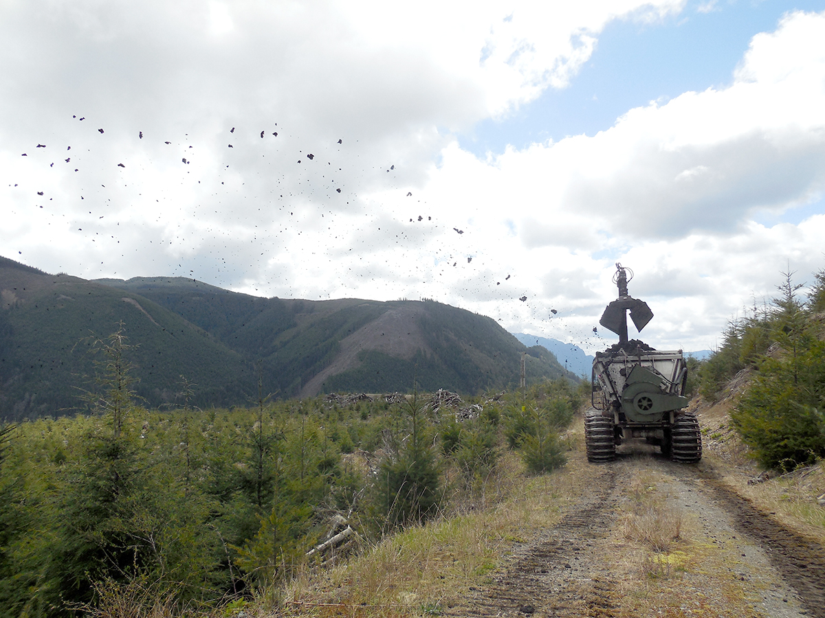 Heavy machinery flings biosolids fertilizer onto a forest land application site, looking over green mountainous terrain.